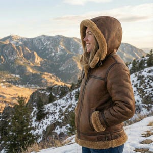 May include: A brown leather jacket with a hood lined with tan fur. The jacket has a zipper closure, pockets, and a belt at the waist. The person is standing on a snowy mountain with a mountain range in the background.