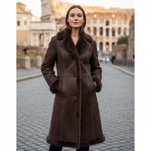 May include: A long, brown shearling coat with a dark brown fur collar and cuffs. The coat has a button closure and pockets. The background features a cobblestone street and a historic building.