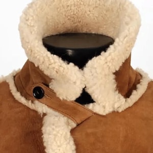 May include: Close-up of a brown suede jacket with a cream-colored shearling collar and lining. The jacket features a high collar, two black buttons, and a leather strap detail. The jacket is displayed on a black mannequin.