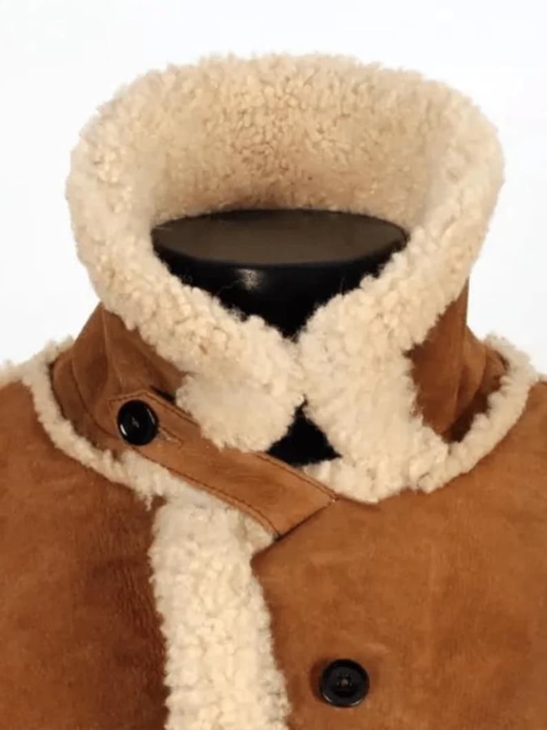 May include: Close-up of a brown suede jacket with a cream-colored shearling collar and lining. The jacket features a high collar, two black buttons, and a leather strap detail. The jacket is displayed on a black mannequin.
