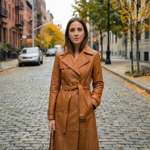 May include: A brown leather trench coat with a double-breasted design, a belt, and pockets. The coat is knee-length and appears to be worn outdoors on a cobblestone street.