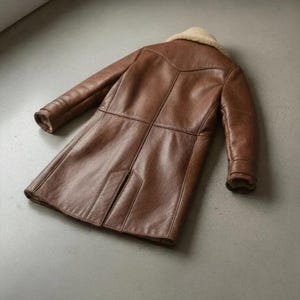 K&ouml;nnte beinhalten: Ein brauner Ledermantel mit einem Shearling-Kragen. Der Mantel ist lang, mit N&auml;hten und Eins&auml;tzen, die einen ma&szlig;geschneiderten Look erzeugen. Die &Auml;rmel sind lang und der Mantel scheint aus einem strapazierf&auml;higen Material zu sein.