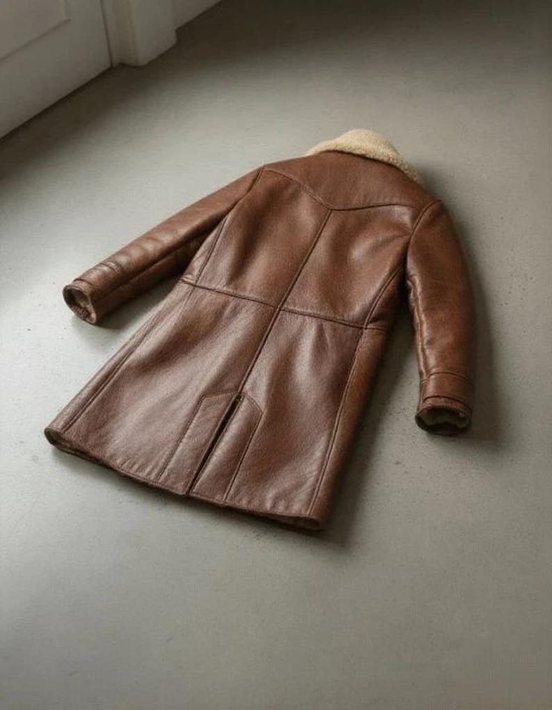 K&ouml;nnte beinhalten: Ein brauner Ledermantel mit einem Shearling-Kragen. Der Mantel ist lang, mit N&auml;hten und Eins&auml;tzen, die einen ma&szlig;geschneiderten Look erzeugen. Die &Auml;rmel sind lang und der Mantel scheint aus einem strapazierf&auml;higen Material zu sein.