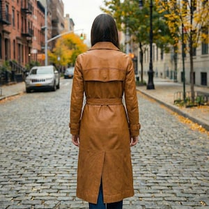 May include: A long, brown leather trench coat with a belted waist and a back vent. The coat has a classic design with a notched collar and buttoned cuffs. The person is standing on a cobblestone street.