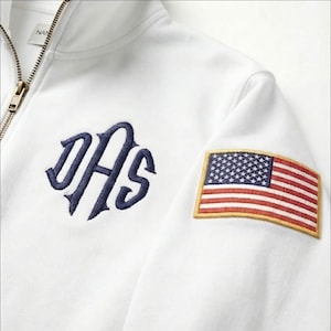 May include: White zip-up sweatshirt with a navy blue monogram and an American flag patch on the sleeve. The sweatshirt has a gold zipper and a tag that says "NAN".