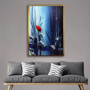 Puede incluir: Pintura floral abstracta en un marco dorado. La obra presenta una amapola roja vibrante sobre un fondo de pinceladas azules y blancas, con elementos altos y oscuros similares a la hierba verde. La pintura se exhibe sobre un sofá gris con cojines decorativos.