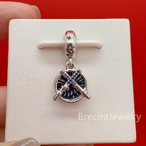 May include: A silver charm featuring a circular pendant with a dark blue background and two crossed lightsabers. One lightsaber is set with pink stones and the other with blue stones. The bail is decorated with small engraved star motifs.
