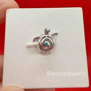 Könnte beinhalten: Ein silberfarbener Schildkröten-Anhänger mit einem spiralförmigen Panzerdesign und kleinen, runden, hellblauen Akzenten am Kopf und in der Mitte des Panzers. Der Anhänger ist auf einer weißen Karte mit dem Aufdruck BreciraJewelry befestigt.