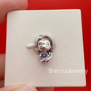 Könnte beinhalten: Ein silberner Charm-Anhänger in Form einer stilisierten weiblichen Figur mit langem Haar, die ein Kleid mit violetten Emaille-Akzenten trägt. Der Anhänger zeigt gravierte Gesichtsdetails und Haarstruktur. Der Text BreciraJewelry steht unten rechts.
