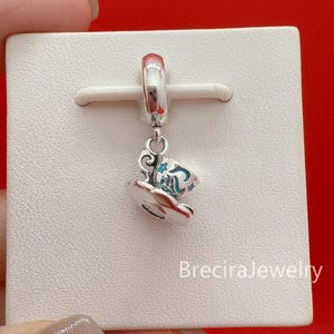 May include: A silver charm featuring a teacup design with blue and white accents. The charm is suspended from a silver loop and is displayed on a white surface. The charm is likely part of a jewelry collection.