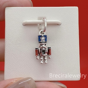 May include: A silver charm in the shape of a toy soldier. The soldier has a blue hat with a star, red arms, and a silver body. The charm is attached to a silver ring. The background is a white card.