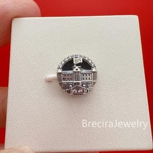 May include: A round silver charm featuring a detailed depiction of a building with a flag. The charm is encrusted with small, clear crystals around the edge. The building has intricate architectural details.