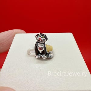 Può includere: Un charm in argento a forma di gatto nero con occhi verdi e naso rosso. Il gatto ha il petto in argento lucido e un piccolo oggetto giallo dietro di sé. Il charm presenta un occhiello in argento per l'aggancio ed è posto su una base bianca.