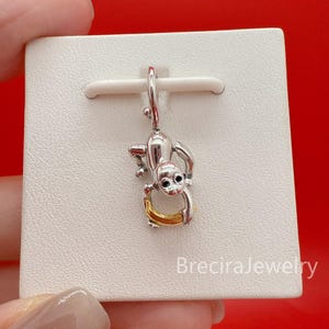 May include: A silver-colored monkey charm holding a gold-colored banana. The charm is hanging from a white card. The monkey has black eyes and is in a playful pose. The charm is likely a pendant or a jewelry component.
