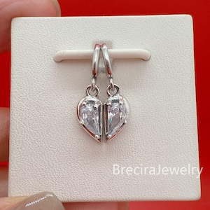 May include: Silver-toned heart-shaped earrings with clear, teardrop-shaped stones. The earrings are displayed on a white card with the text "BreciraJewelry" in the bottom right corner. The background is a solid red.