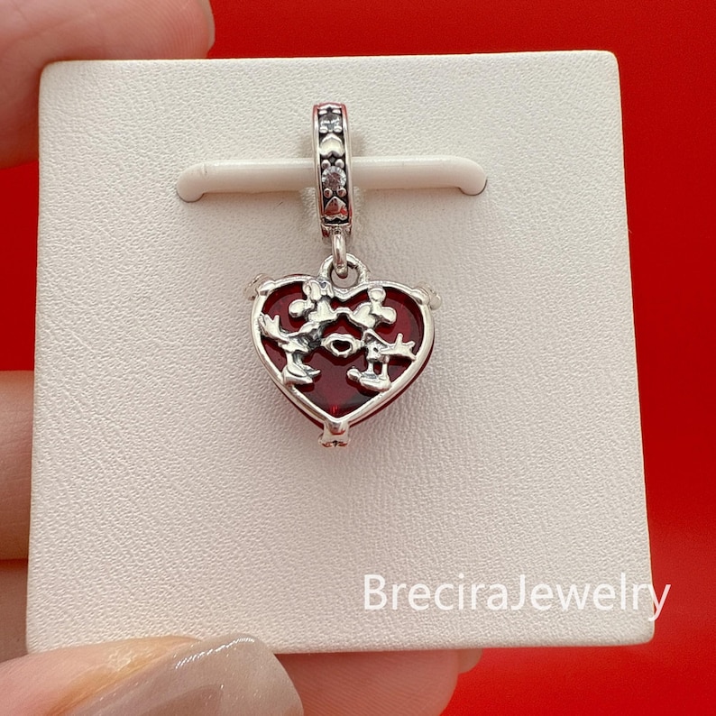 Puede incluir: Un charm de plata con un colgante rojo en forma de coraz&oacute;n con siluetas de Mickey y Minnie Mouse. El charm tiene un peque&ntilde;o lazo con detalles decorativos y se muestra sobre una superficie blanca sobre un fondo rojo.