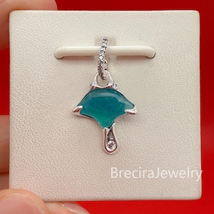 Op de afbeelding: Een zilveren hanger in de vorm van een mantarog met een doorschijnend blauwgroen lichaam, voorzien van een oogje en een klein accentpunt bezet met heldere steentjes. De hanger wordt getoond op een vierkante kaart met de tekst BreciraJewelry.