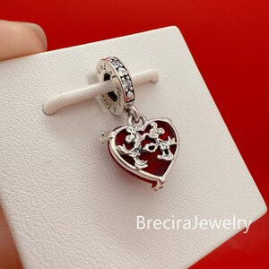 Puede incluir: Un charm de plata con un colgante en forma de coraz&oacute;n rojo. El coraz&oacute;n tiene un dise&ntilde;o decorativo y est&aacute; unido a una banda plateada con las palabras "kind" y "love". El charm se muestra sobre una superficie blanca sobre un fondo rojo.