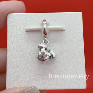 May include: A silver dangle charm featuring a stylized character head with a circular cutout in the center, suspended from a polished silver bail. The charm is displayed on a white square card with the text BreciraJewelry printed in the bottom corner.