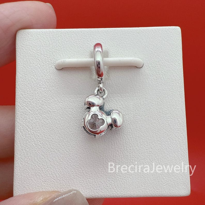 May include: A silver dangle charm featuring a stylized character head with a circular cutout in the center, suspended from a polished silver bail. The charm is displayed on a white square card with the text BreciraJewelry printed in the bottom corner.