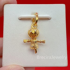 Può includere: Un ciondolo color oro a forma di spaventapasseri con corpo testurizzato, braccia tese e testa rotonda. La figura presenta piccole pietre chiare incastonate negli occhi e sul busto, sospesa a un occhiello circolare color oro.