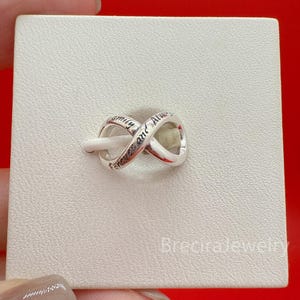 Puede incluir: Un charm de infinito en tono plateado con el texto grabado "Family and Always" a lo largo de su superficie curva. El charm está montado sobre una tarjeta de presentación cuadrada de color blanco roto con textura. El metal tiene acabado pulido.