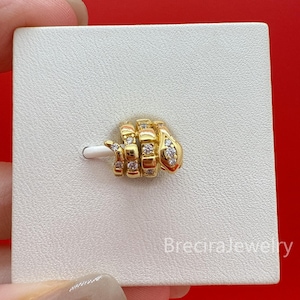 May include: A gold-colored ring in the shape of a hand making a gesture. The ring is adorned with small, clear gemstones. The ring is on a white card, with the text "BreciraJewelry" in the bottom right corner.