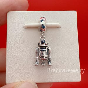 May include: A silver charm shaped like the R2-D2 droid, featuring blue and black enamel detailing on the body and head. The bail is decorated with small blue star-shaped accents. The charm is displayed on a white square card with the text BreciraJewelry.