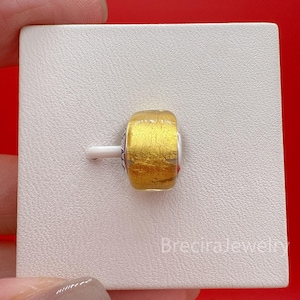 Op de afbeelding: Een cilindervormige bedel met een textuur van goudkleurige metaalfolie ingesloten in helder glas. De bedel is bevestigd op een witte plastic displaypen. De tekst BreciraJewelry staat rechtsonder op de display gedrukt.