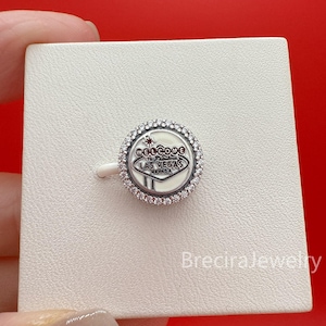 Puede incluir: Un charm de plata con un fondo blanco con el texto "Welcome to Fabulous Las Vegas Nevada". El charm redondo tiene un borde de pequeñas piedras transparentes. El charm está sobre un fondo blanco.