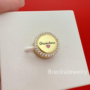 Pode incluir: Um berloque dourado com a palavra "Grandma" em escrita preta e um coração rosa. O berloque é rodeado por pequenos cristais transparentes. O berloque está sobre uma superfície branca.