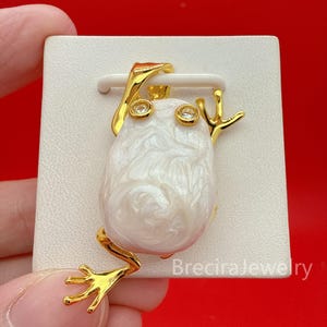 May include: A gold-toned metal frog pendant featuring a large, irregular white baroque pearl as the body. The frog has two small, clear round stones set as eyes and a polished gold finish on the limbs and head. The text BreciraJewelry is visible.