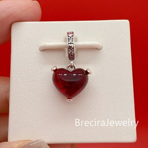 Puede incluir: Un charm de plata con un colgante en forma de coraz&oacute;n rojo. El coraz&oacute;n tiene una superficie texturizada y est&aacute; suspendido de una fianza decorativa de plata. El charm se muestra en una tarjeta blanca sobre un fondo rojo. El texto "BreciraJewelry" es visible.