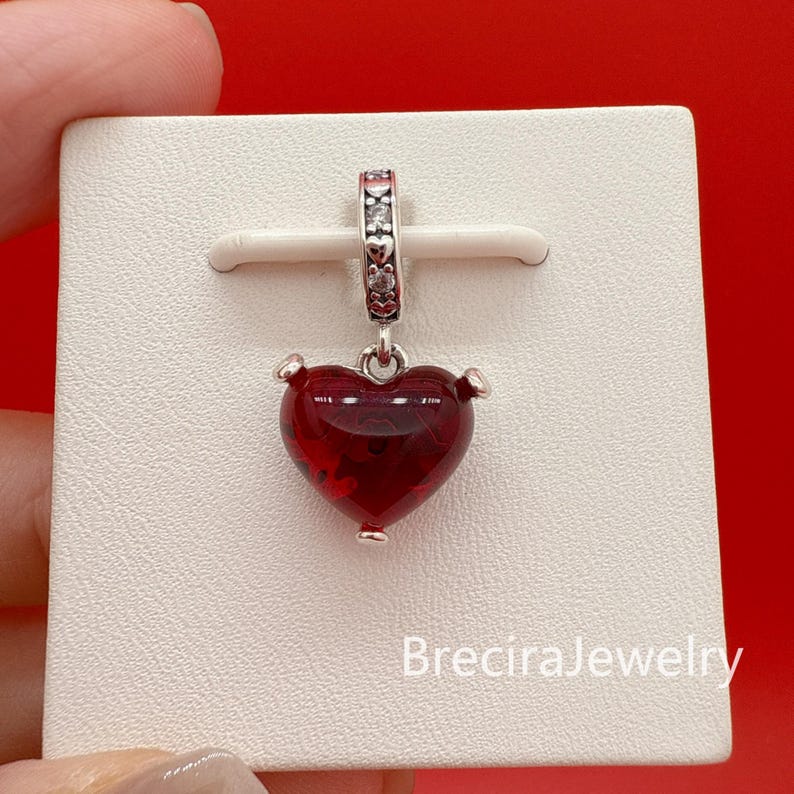 Puede incluir: Un charm de plata con un colgante en forma de coraz&oacute;n rojo. El coraz&oacute;n tiene una superficie texturizada y est&aacute; suspendido de una fianza decorativa de plata. El charm se muestra en una tarjeta blanca sobre un fondo rojo. El texto "BreciraJewelry" es visible.