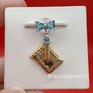 Op de afbeelding: Een zilverkleurige bedel met een blauwe kristallen strik aan de bovenkant, verbonden aan een goudkleurige notitieboek- en potloodhanger. Het notitieboek heeft een spiraalbinding en een kleine blauwe steen. De tekst BreciraJewelry is onderaan zichtbaar.