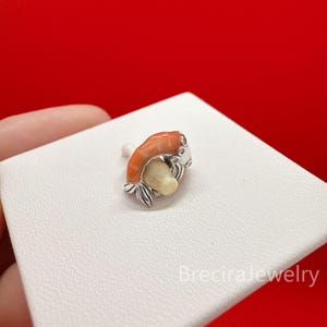 Könnte beinhalten: Ein Silberring mit einem Pilzmotiv, das einen leuchtend orangefarbenen Hut und einen cremefarbenen Stiel aufweist. Die Metallbasis des Rings ist unten mit kleinen silbernen Blättern verziert. Der Text BreciraJewelry ist unten rechts sichtbar.