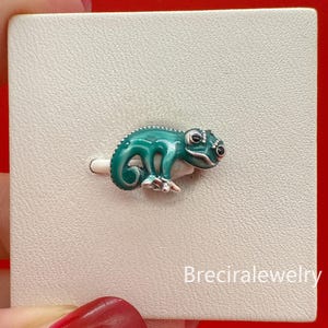 May include: A teal enamel chameleon brooch with silver accents. The lizard-shaped pin has black eyes and is set against a white background. The jewelry is a unique accessory.