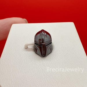Puede incluir: Un pendiente de botón con forma de casco en gris metálico oscuro con una abertura en forma de T para la visera. La pieza tiene una superficie lisa y reflectante y un pequeño poste blanco en el lateral. El texto BreciraJewelry aparece abajo.