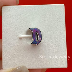 Op de afbeelding: Een zilveren ring met een letter D-initiaal met een paarse en blauwe iriserende inleg. De letter is bevestigd op een witte vierkante presentatiekaart met de tekst BreciraJewelry gedrukt in de rechterbenedenhoek van het kaartoppervlak.