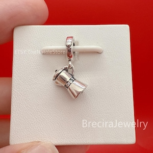 Op de afbeelding: Een zilverkleurige bedel in de vorm van een koffiezetapparaat, met een kleine lus om aan een armband of ketting te bevestigen. De bedel heeft een gedetailleerd ontwerp, met een handvat en een decoratieve band. Gepresenteerd op een witte ondergrond.