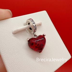 Puede incluir: Un charm en forma de coraz&oacute;n con un centro rojo transl&uacute;cido y detalles plateados. El charm est&aacute; suspendido de un anillo plateado con piedras transparentes y la palabra "Pandora" grabada. El charm se muestra sobre una superficie blanca sobre un fondo rojo.