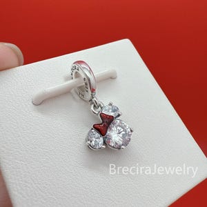 May include: A silver charm featuring a clear, round, faceted cubic zirconia stone and a smaller clear stone, accented with a small, glossy red enamel bow. The charm is engraved with the text PANDORA S925 ALE on the circular bail.