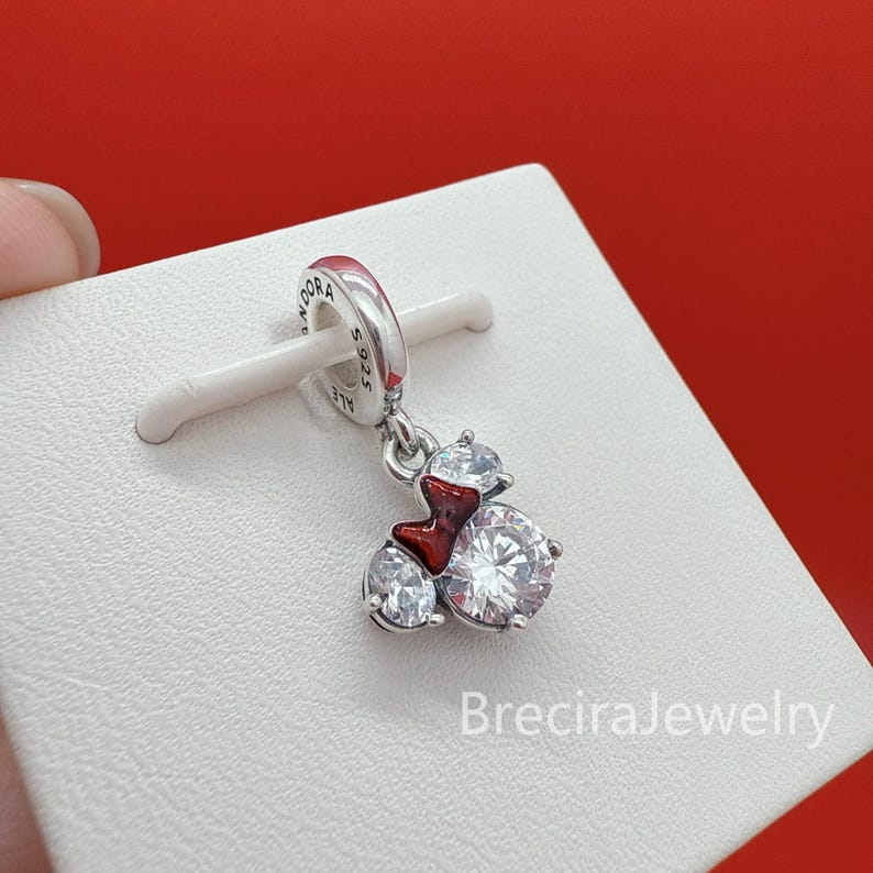 May include: A silver charm featuring a clear, round, faceted cubic zirconia stone and a smaller clear stone, accented with a small, glossy red enamel bow. The charm is engraved with the text PANDORA S925 ALE on the circular bail.
