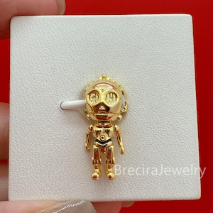 May include: A gold-toned metal charm shaped like a robotic character with a rounded head, large circular eyes, and a segmented body. The charm is attached to a white display card featuring the text BreciraJewelry printed in the bottom right corner.