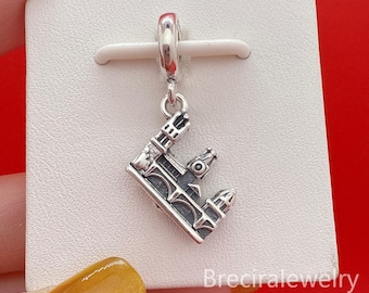 May include: A silver-colored charm in the shape of a detailed bridge, likely a jewelry piece. The charm hangs from a silver loop, suggesting it's designed to be added to a bracelet or necklace. The bridge design features architectural details.