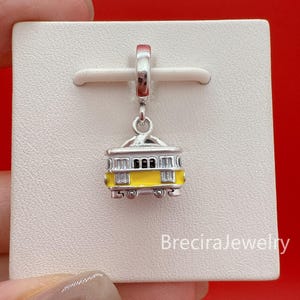Può includere: Un charm d'argento raffigurante un tram in miniatura giallo e argento. Il tram ha finestre nere ed è sospeso a un'ansa d'argento. Il charm è presentato su uno sfondo bianco con uno sfondo rosso.