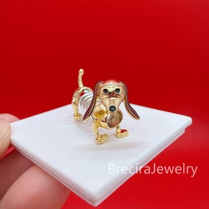 Peut inclure: Une figurine de teckel dorée avec un corps à ressort, de longues oreilles tombantes aux accents marron foncé, ainsi que des yeux et un nez en émail noir. Le chien a une finition métallique et repose sur une base carrée blanche avec le texte BreciraJewelry.