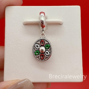 Könnte beinhalten: Ein silberner Charm-Anhänger mit einem kreisförmigen Anhänger mit roten, grünen und weißen Emaille-Akzenten. Der Charm hat ein dekoratives Muster am Rand und eine kleine Silberschlaufe oben zur Befestigung. Der Charm wird auf einer weißen Oberfläche präsentiert.