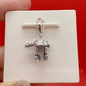 May include: A silver-toned metal charm shaped like a four-legged walker vehicle. The charm features intricate mechanical detailing and is suspended from a small bail decorated with engraved star patterns, attached to a white display card.