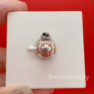 May include: A silver BB-8 charm with orange and red detailing. The charm is a miniature replica of the Star Wars droid, featuring detailed panels and a circular design. The charm is displayed on a white surface against a red background.